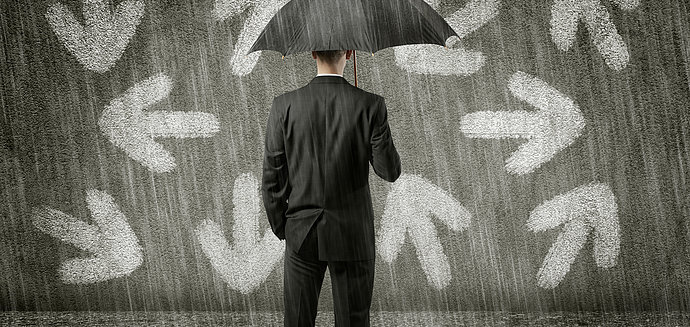 Mann mit Schirm steht ratlos vor einer Wand mit Pfeilen in verschiedene Richtungen