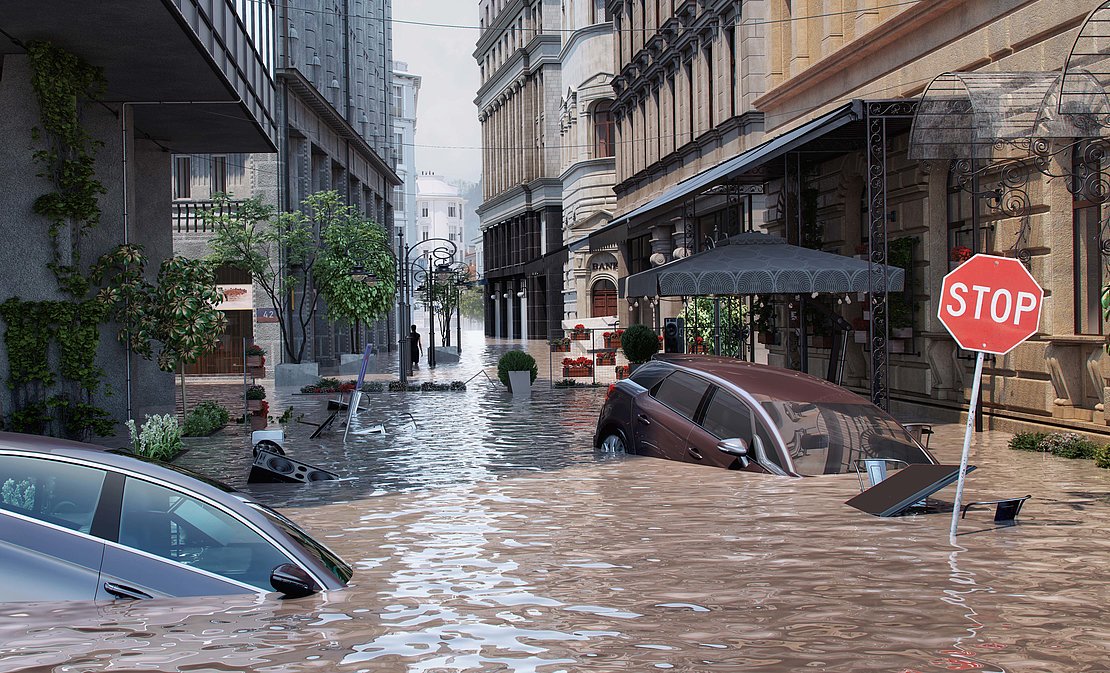Überflutete Straße mit 2 PKW, die bis zur Motorhaube im Wasser stehen. Rechts und links gesäumt von Gebäuden.